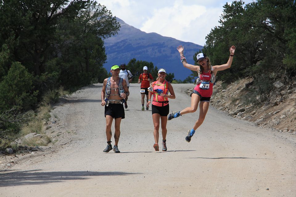 Heather Hart, ultramarathon runner,  leaping through the air as she runs with two other runners running down a dirt road with mountains in the background 