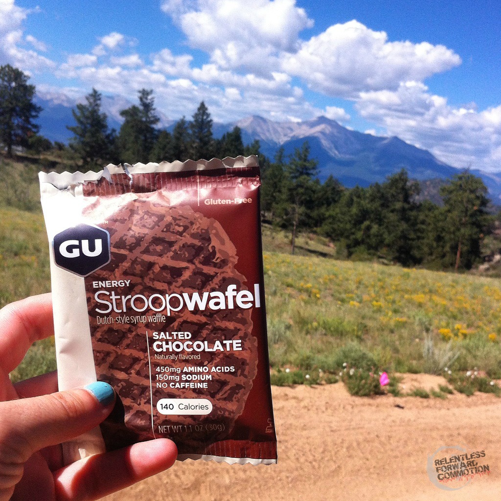 A hand holding a GU stroopwafel on a trail with mountains and fields in the background