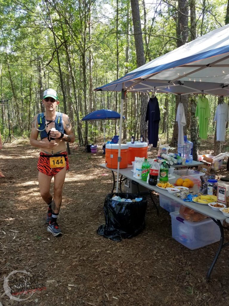 runner stands in front of an ultramarathon fueling aid station, covered in food and drinks for ultrarunners to eat during the race