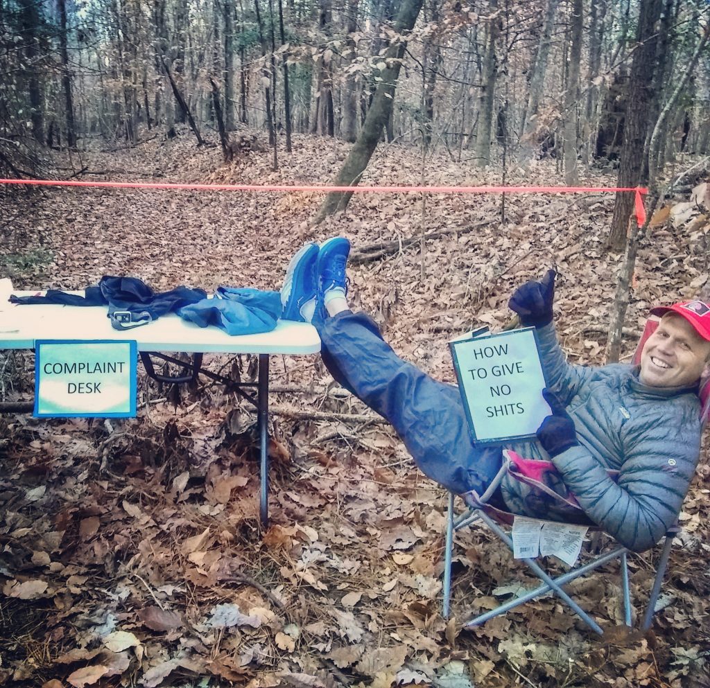 Image of a volunteer at an ultramarathon with his feet up on a table, sitting in a chair, with the sign "complaint desk" on the table, and holding a notebook that says "how to give no shits"