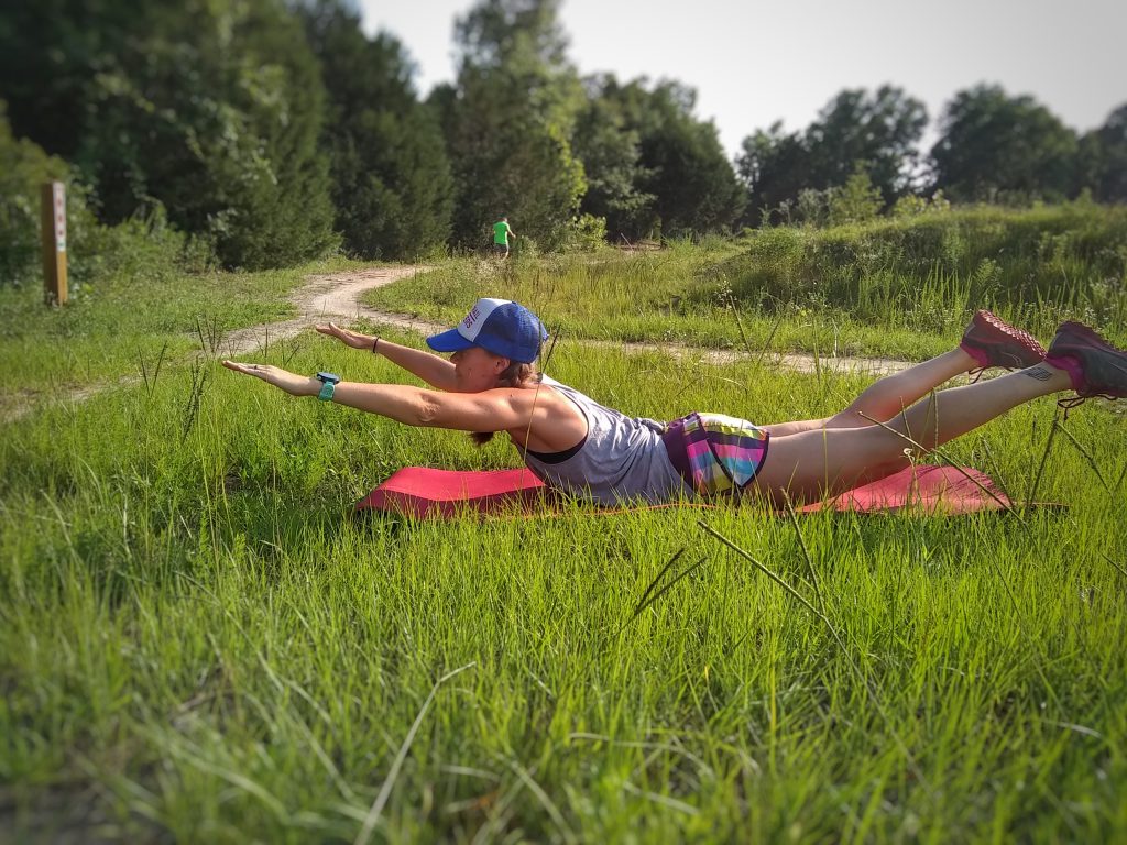 Ultramarathon runner demonstrating bodyweight strength training exercises in the form of a Superman low back extension while lying on a yoga mat next to a trail