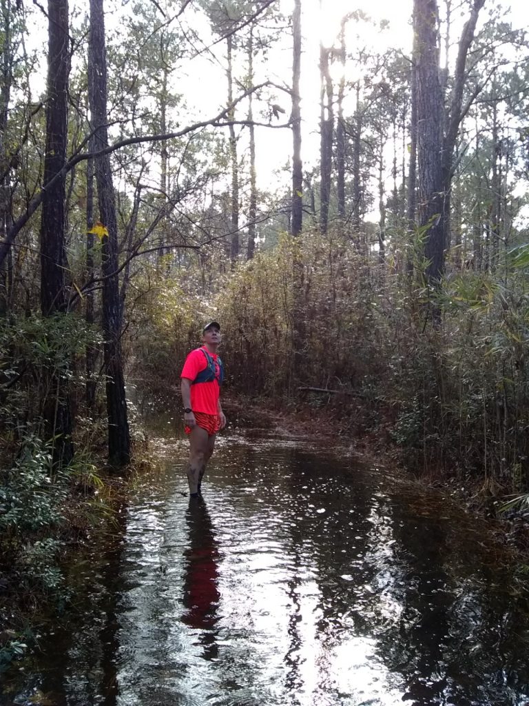Image of Geoff Hart standing in the middle of a flooded forest trail, with water coming up and over his ankles. 