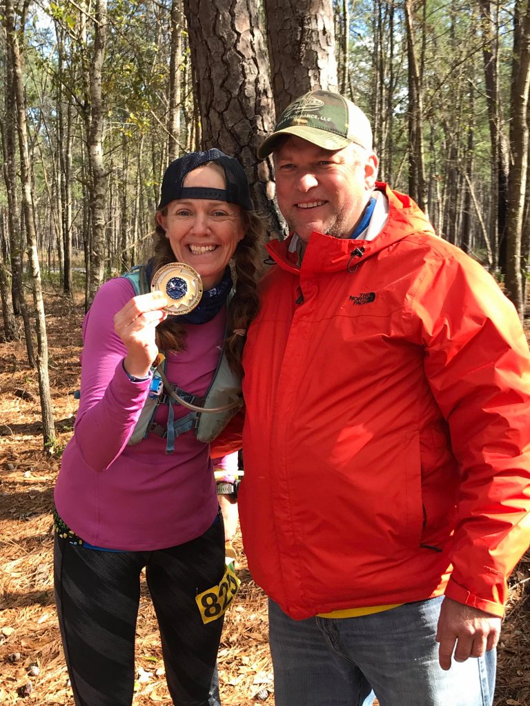 Heather Hart holding a finishers belt buckle from a 100 mile ultramarathon , hugging race director Chad Haffa at the finish line
