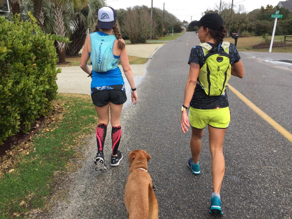 Image of the back of two runners running down the pavement, wearing hydration packs and running with a dog. 