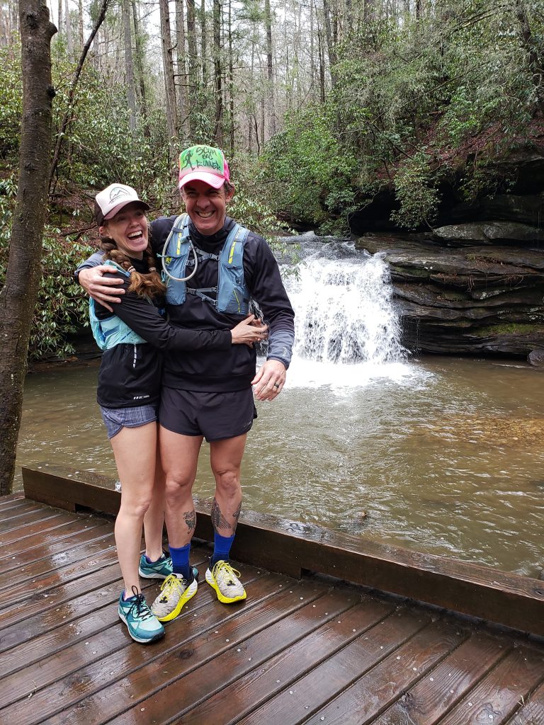 Ultramarathon coaches Heather and Geoffrey Hart standing on a wooden walkway in front of a waterfall