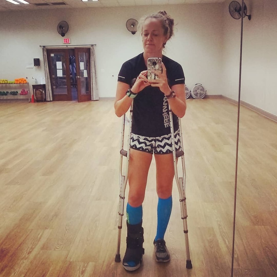 An injured runner takes a picture in a mirror of a gym fitness studio, wearing a recovery boot on one leg, and resting on crutches