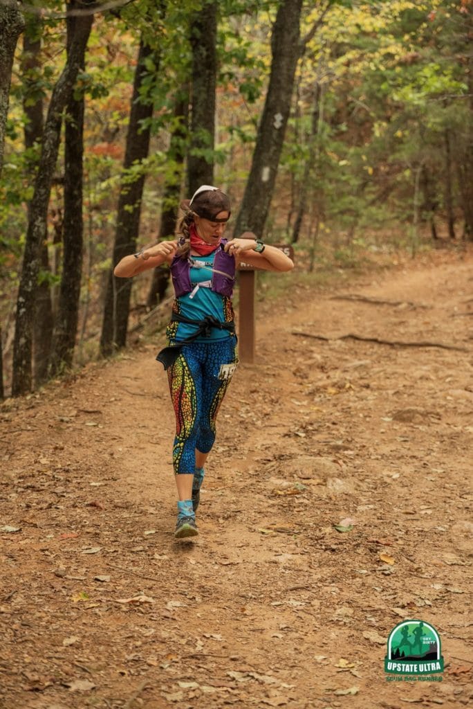 Tired, sore, ridiculous, hungry ultrarunner running down a trail in the woods looking for food in her pockets, oblivious to the camera pointed right at her. 