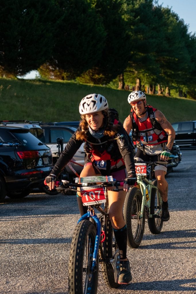 Image of a smiling woman on a mountain bike with a man on a bike behind her finishing up an Adventure Race 