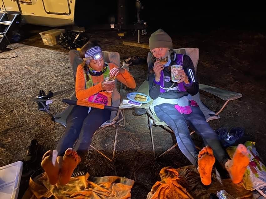 Two ultrarunners eating food and warming their feet by the fire during the overnight portion of a 100 mile race