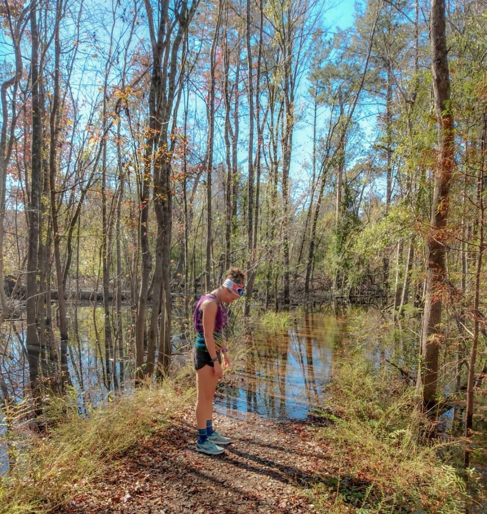 Heather Hart, stopped on the middle of a trail that is flooded over, making a sad face over not wearing waterproof trail running shoes. 
