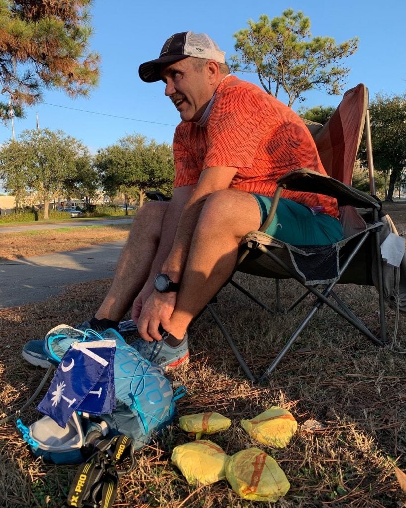 Image of an ultrarunner sitting in chair tying shoes, hydration pack and four McDonalds cheeseburgers laying on the ground in front of him. 