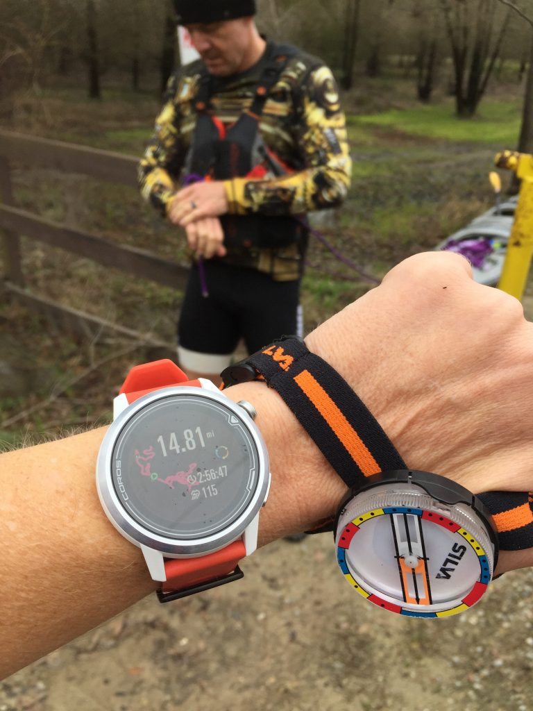 Image of a COROS APEX 46mm on a wrist, with a SILVA compass also on the wrist, with man in the background wearing a life jacket while adventure race training