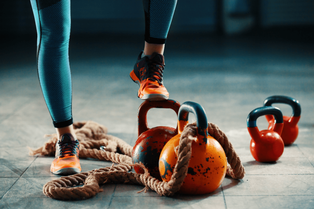 Women's foot in running sneakers on a kettlebell with multiple kettlebells and a fitness rope in the background.