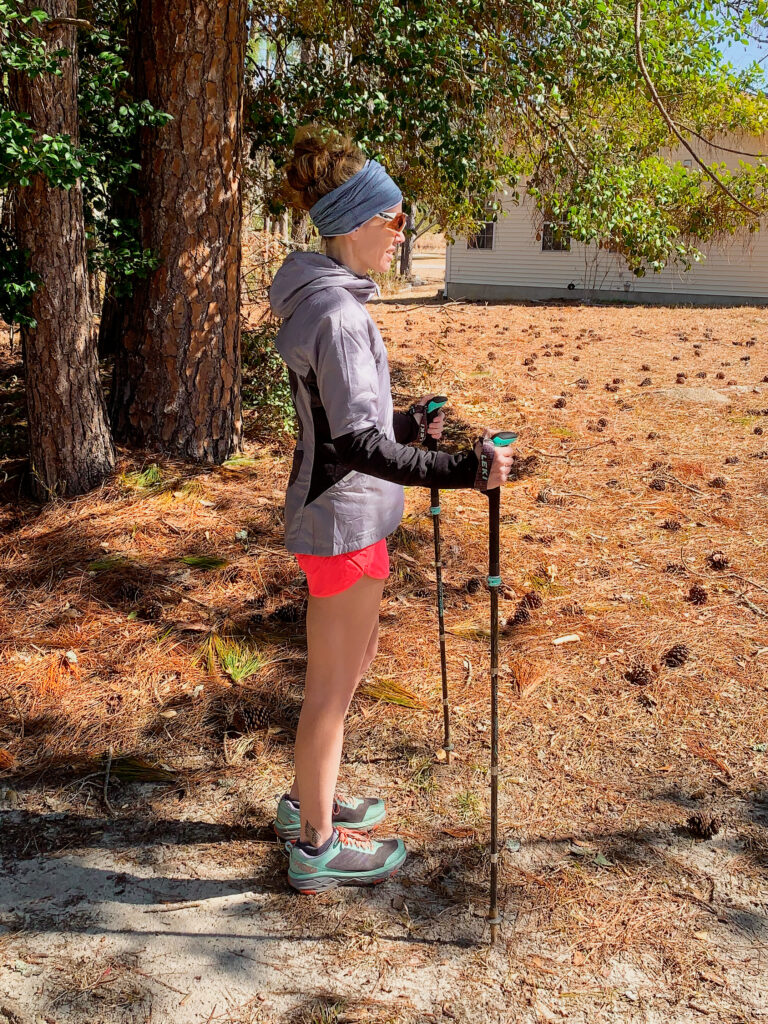 Heather Hart demonstrating the ideal fit for trekking poles: Ideally, when standing on flat ground, you can hold your arms at your sides, elbows bent at a 90-degree angle, with your hands on the trekking pole grips, and the pole tips on the ground. 