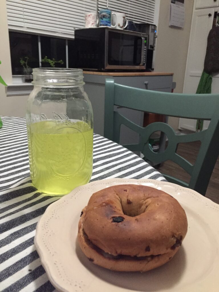Image of a bagel on a plate with a drink in a glass next to it, on a kitchen table 