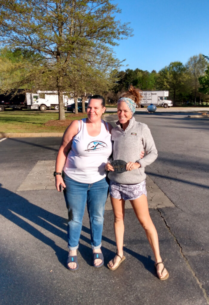 Holly Shoemaker and Heather Hart celebrating post Country Mile 100 mile ultramarathon, holding a belt buckle