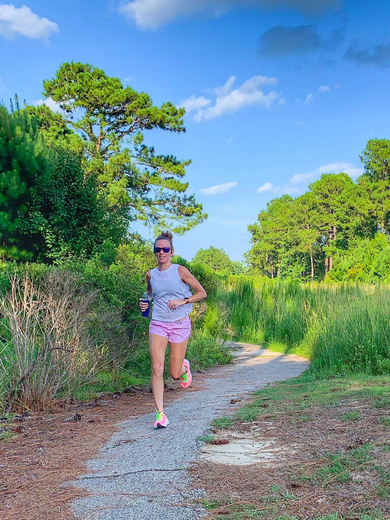 Heather Hart running down a paved golf cart path in an overgrown abandoned golf course, blue sky and very green grass