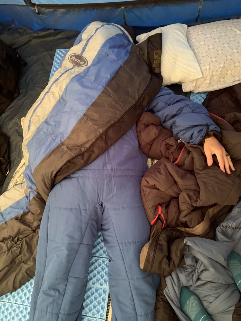 Man in a selk'bag sleeping bag suit covered in various sleeping bags asleep in a tent