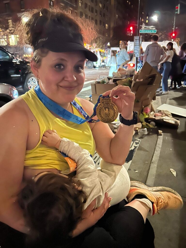 Marathon Mom Dana proudly showing off her NYC marathon medal while breastfeeding her baby at the finish line after running the NYC marathon. 