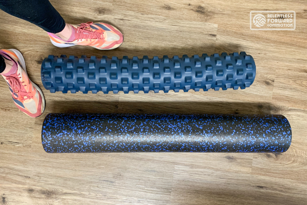 Two foam rollers on the floor pictured next to a runner's feet in running shoes