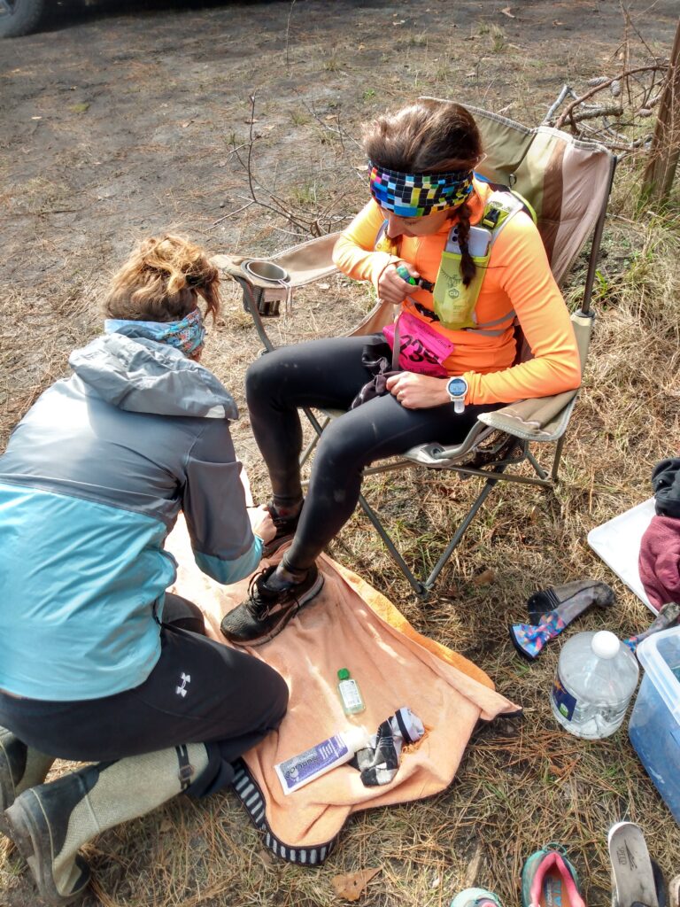Image of an ultrarunner sitting in a chair while a crew member tends to her feet during a race 